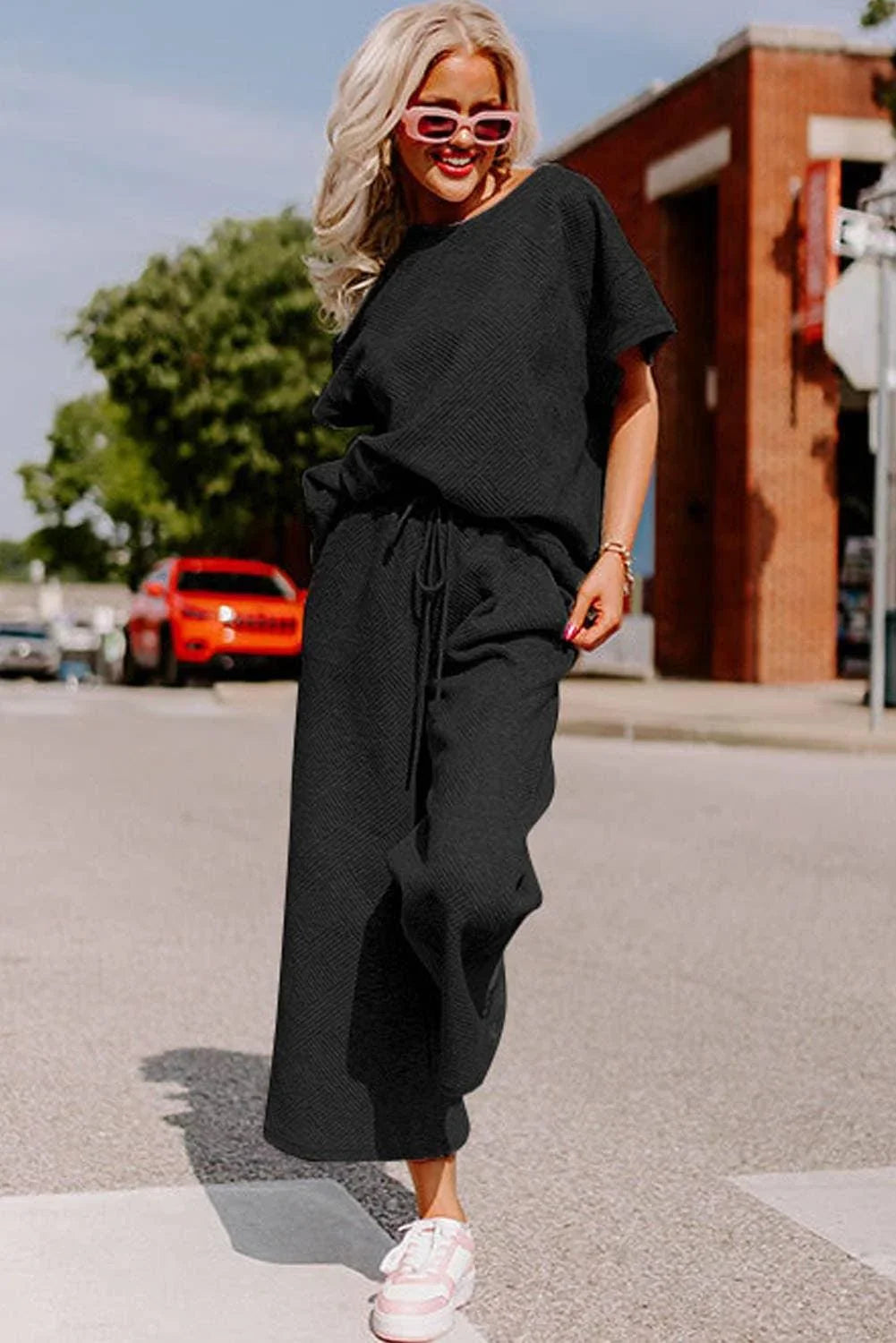 Woman in black casual matching set, pink sunglasses, and sneakers walking on city street