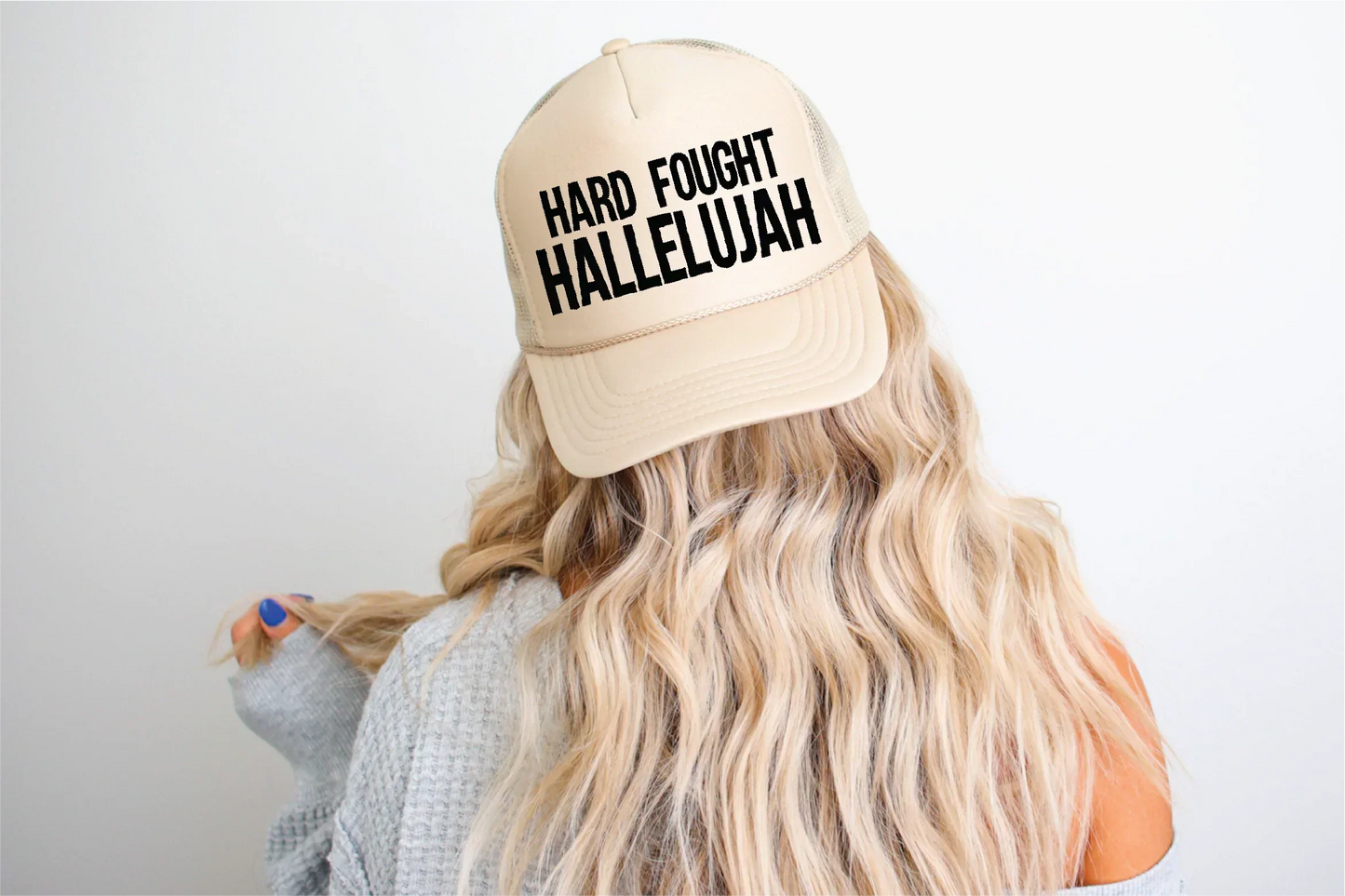 Beige trucker hat with 'HARD FOUGHT HALLELUJAH' text, worn by woman with wavy blonde hair