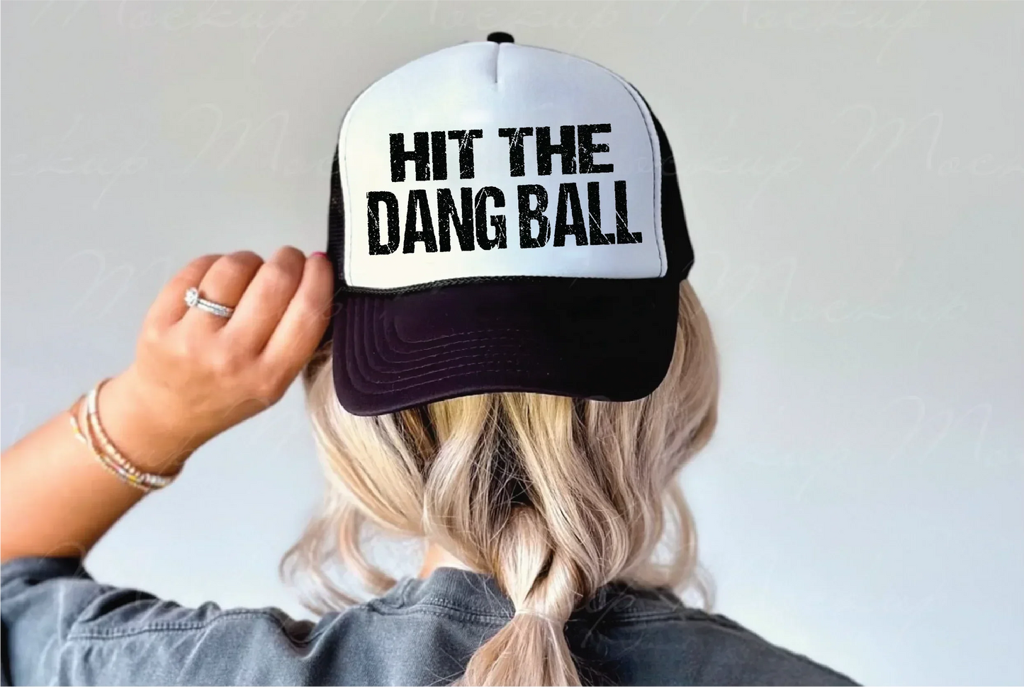 White and black trucker hat with 'HIT THE DANG BALL' text, worn by blonde woman with braid