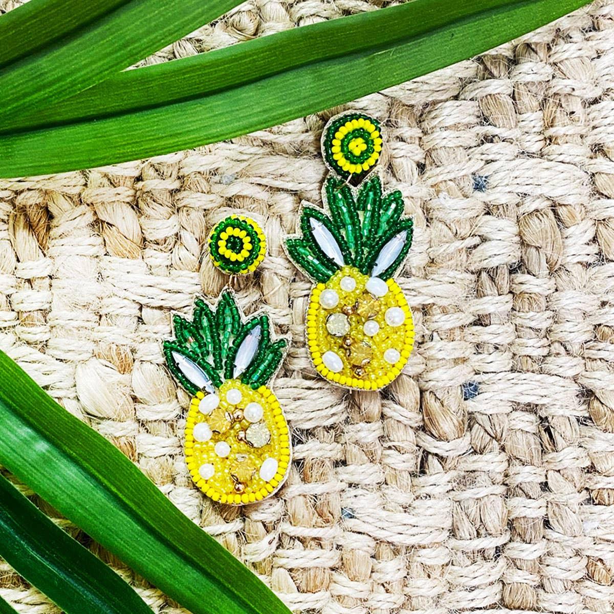 Beaded pineapple earrings with green leaves and yellow body, styled on woven mat with tropical leaves