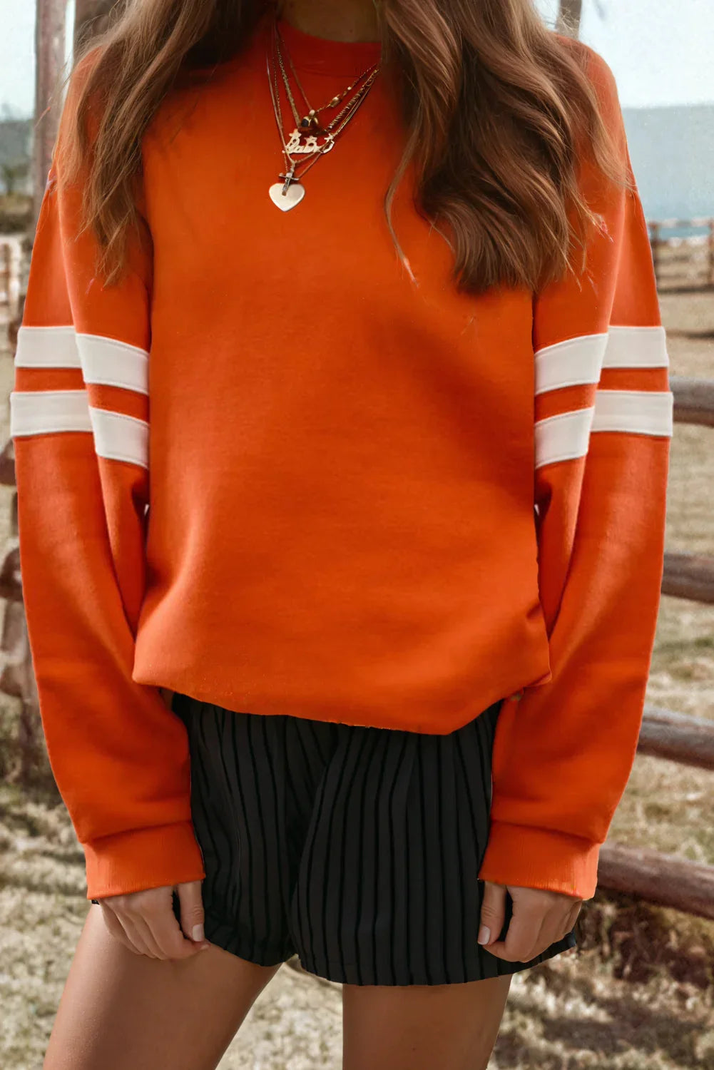 Orange striped sleeve sweater with layered necklaces and black pleated shorts