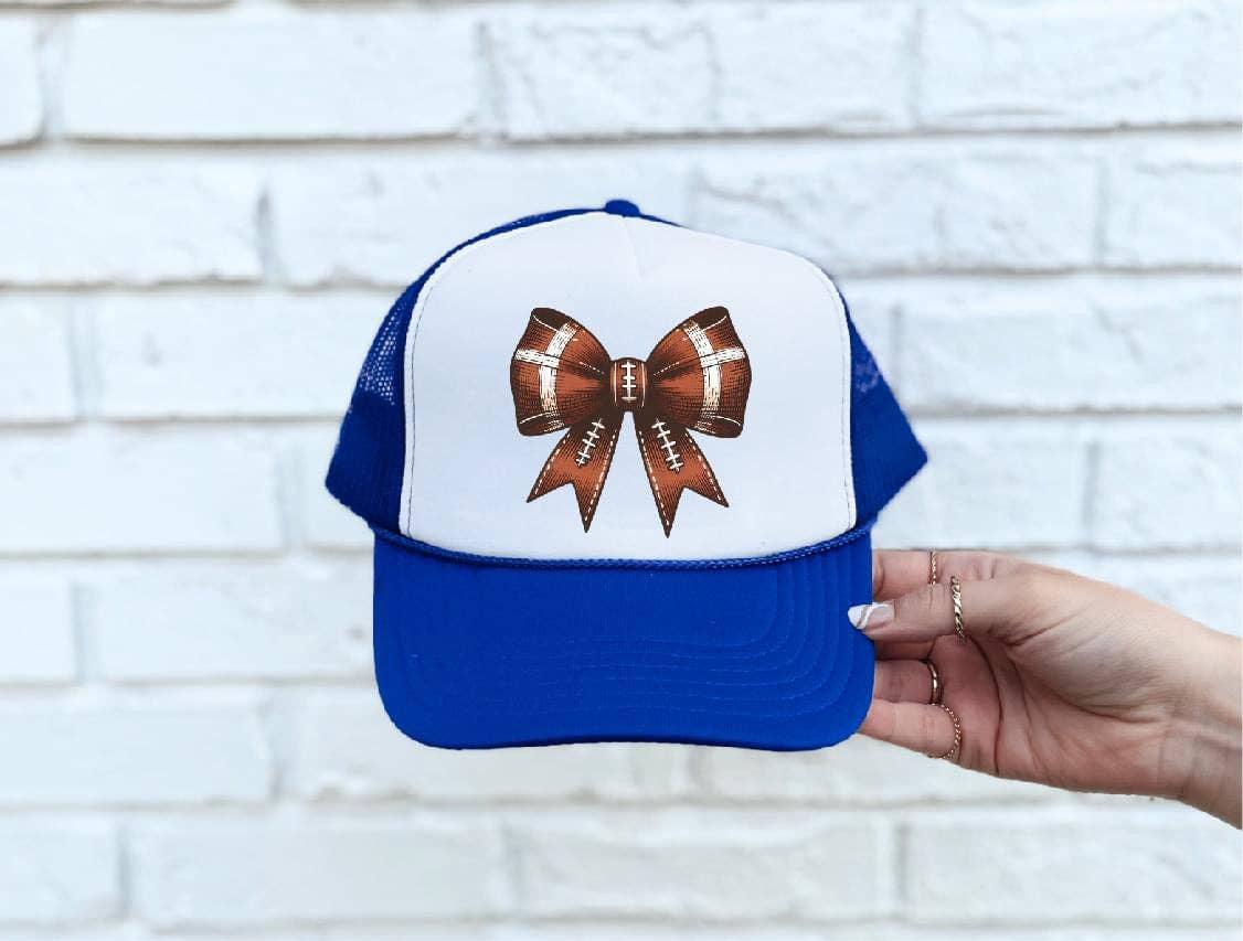Blue and white trucker hat with football bow design, held against white brick wall