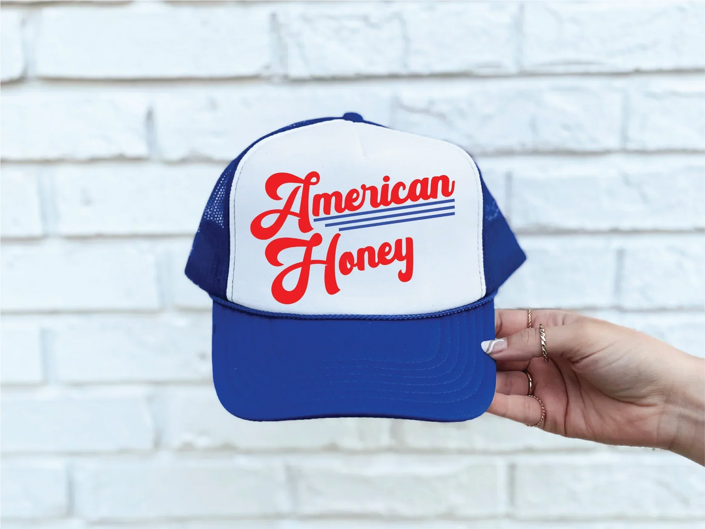Blue and white trucker hat with 'American Honey' in red script, held against white brick wall