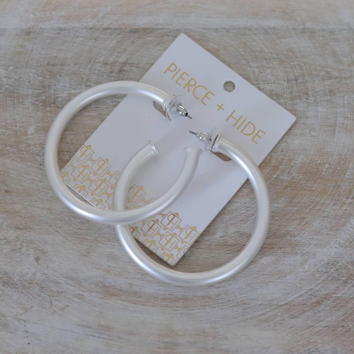 Pair of large matte silver hoop earrings on a Pierce + Hide card, on wood background