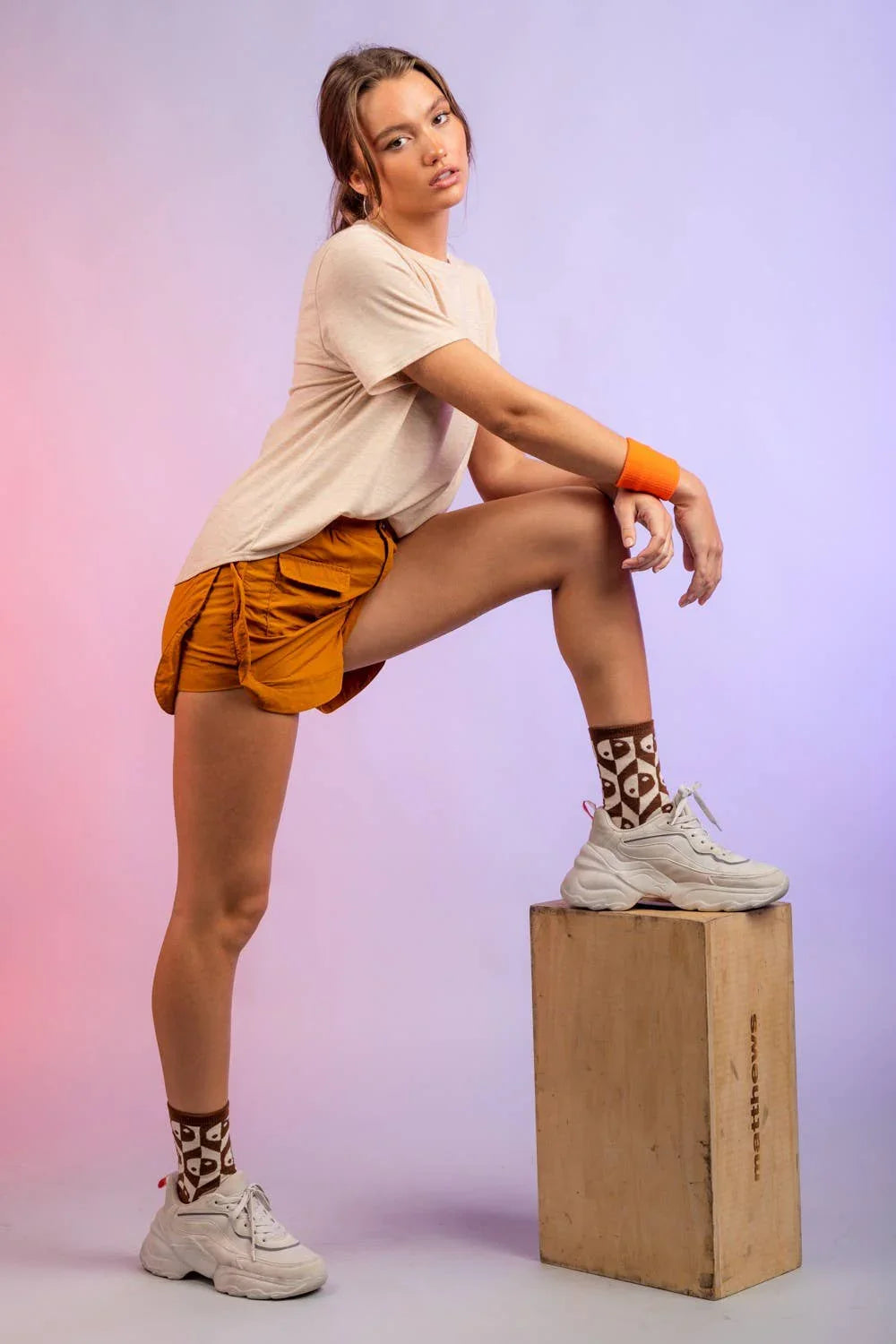 Woman in beige t-shirt and orange shorts posing with one leg on a wooden box, wearing chunky sneakers and patterned socks, athletic retro style