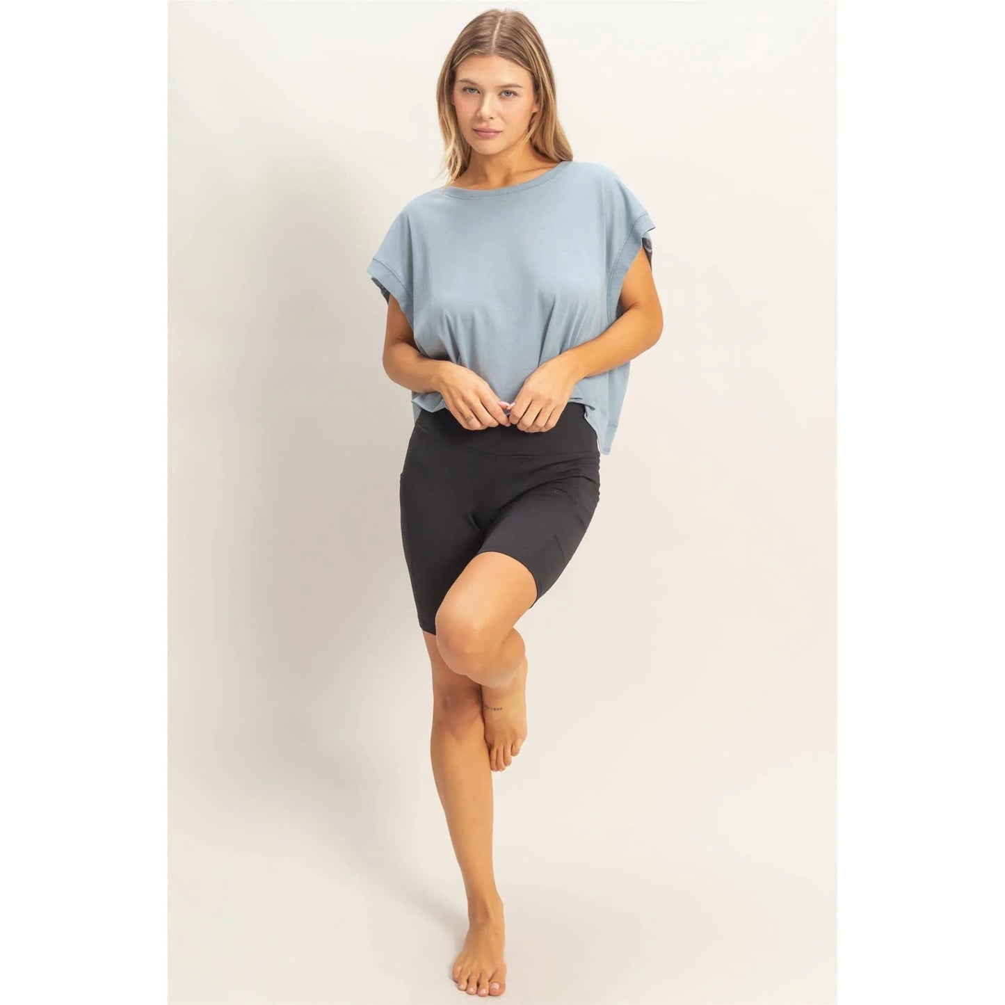 Woman in blue loose t-shirt and black biker shorts posing barefoot against a plain background