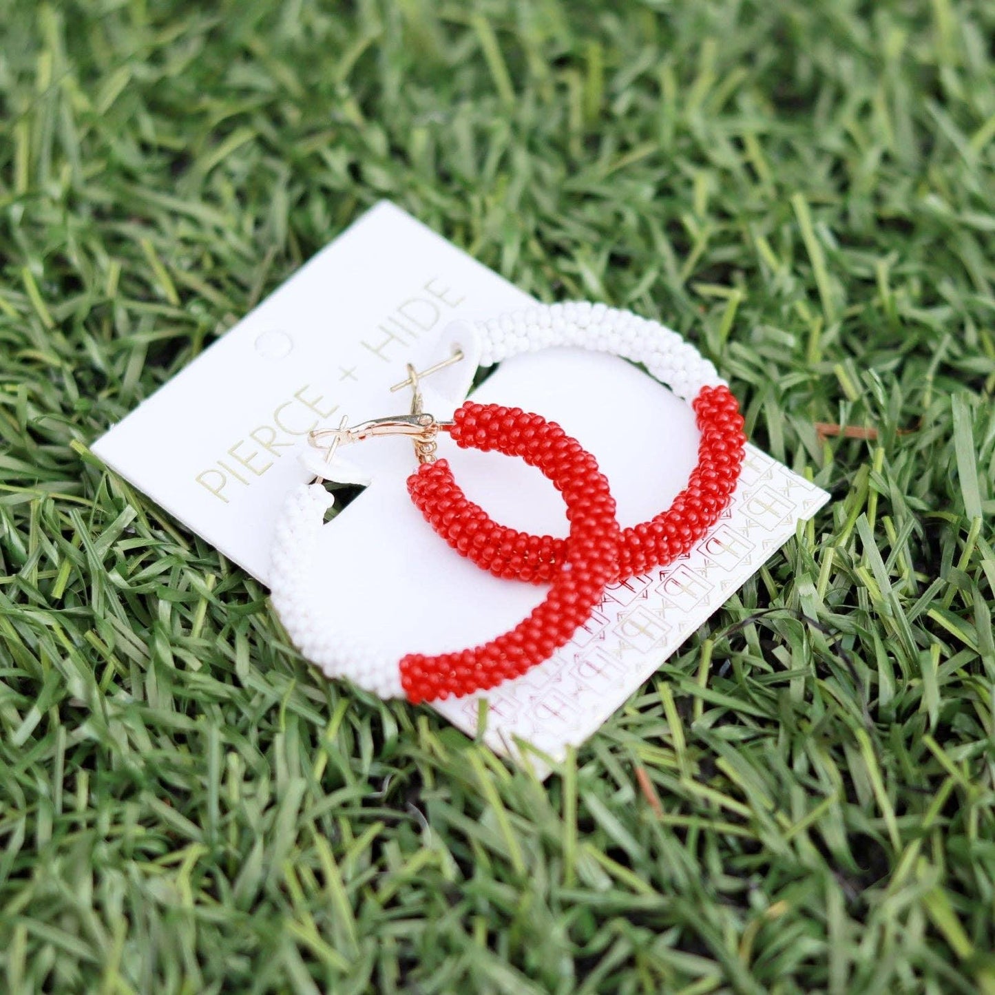 Red and white beaded hoop earrings on branded card displayed on green grass