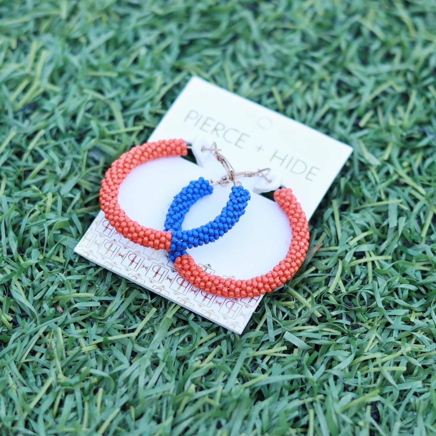 Beaded hoop earrings in orange and blue on a display card, placed on green grass