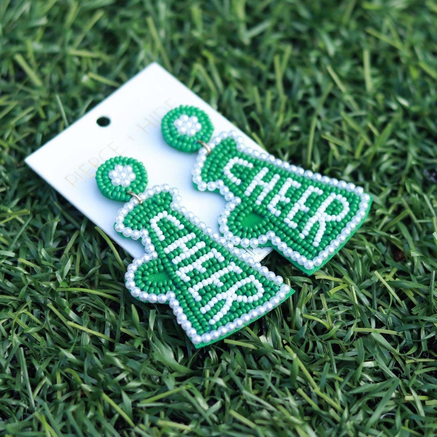 Green beaded cheer megaphone earrings with white accents on display card, on grass