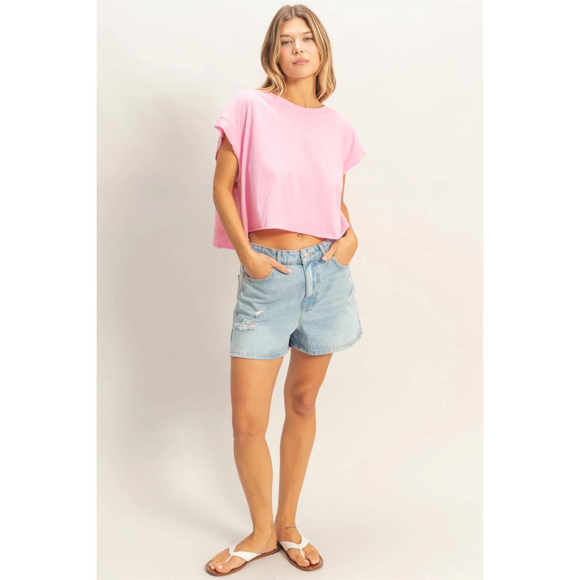 Woman wearing a pink crop top, light denim distressed shorts, and white sandals