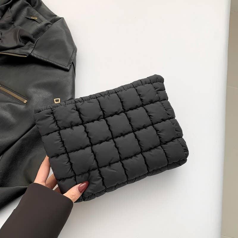 Hand holding a black quilted clutch purse next to a black leather jacket on a white surface.