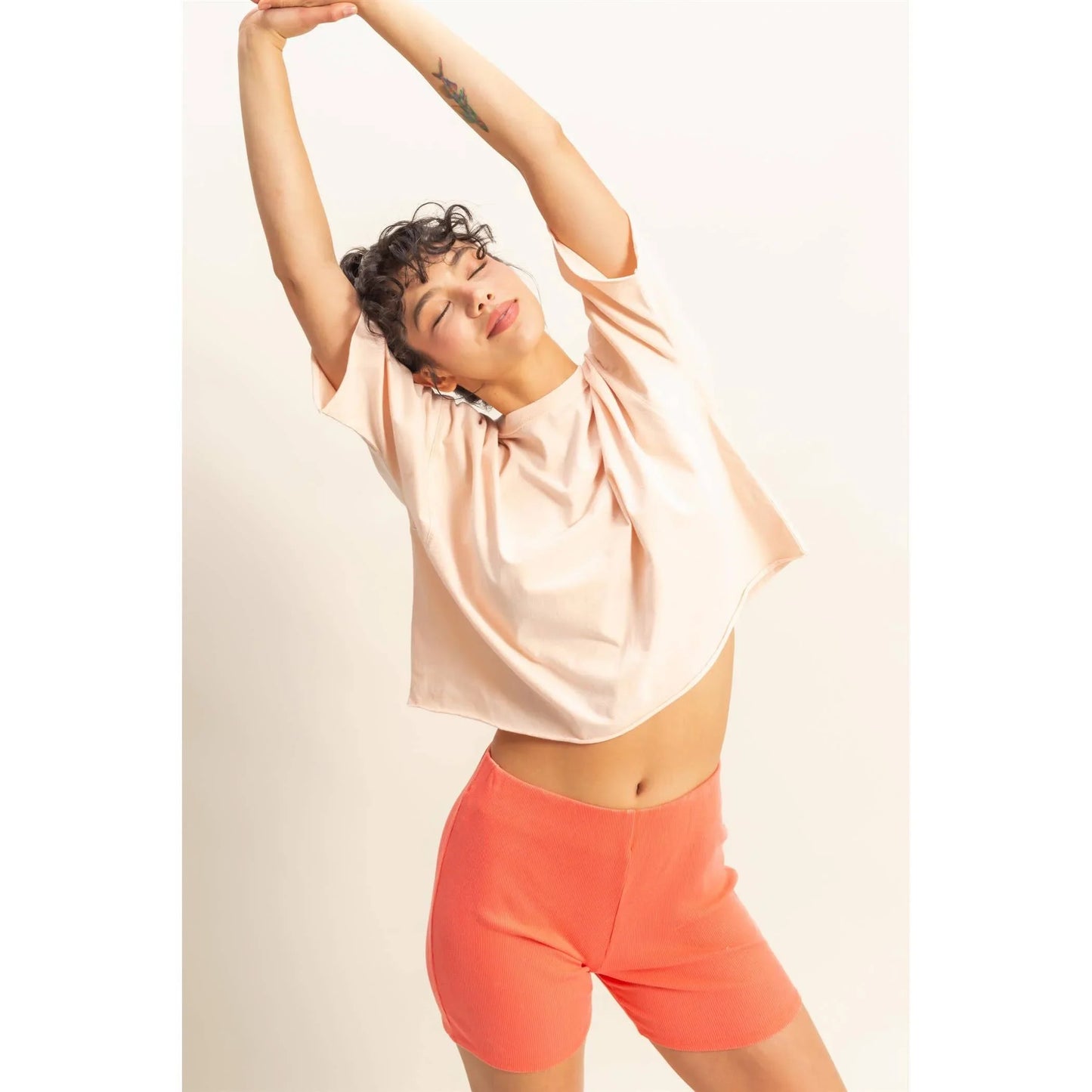 Woman in peach crop top and coral shorts stretching arms overhead, stylish casual outfit