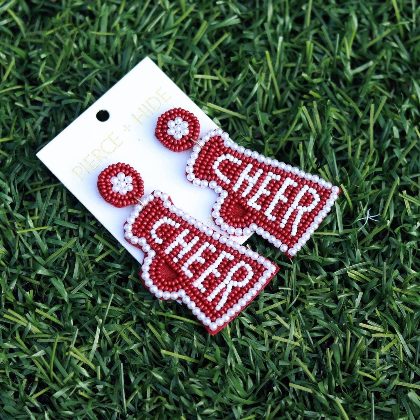 Red beaded CHEER megaphone earrings on display card, styled on green artificial grass.